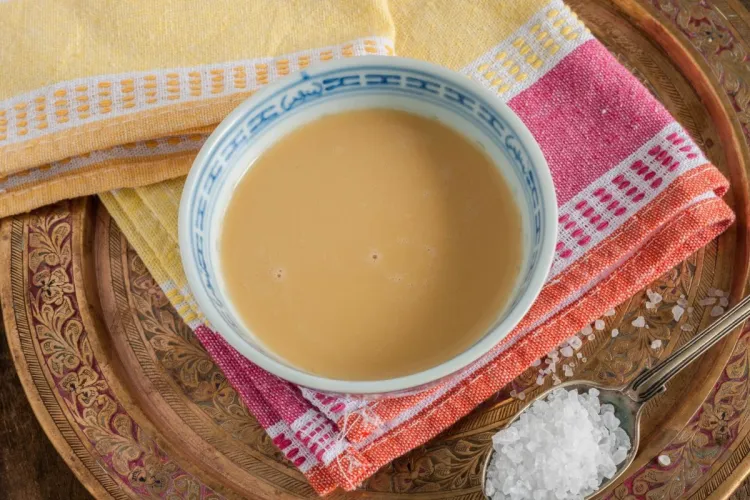 Cuenco de té tibetano con mantequilla, de color cremoso, sobre un paño de rayas y una bandeja de metal labrado, con una cucharilla de sal gorda al lado.