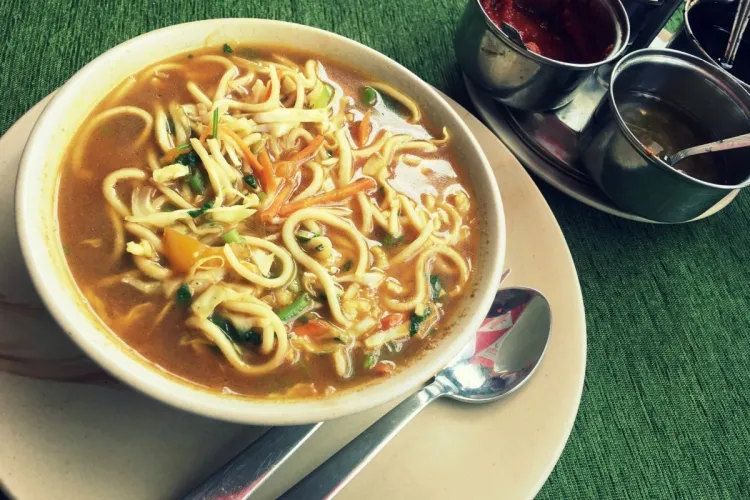 Cuenco de thukpa, sopa tibetana de fideos con verduras en un caldo especiado, con cuencos de salsa al lado y una cuchara.