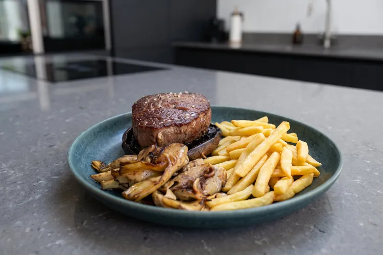 Tournedó de solomillo marcado sobre un sombrero de portobello, con alcachofas y setas salteadas y patatas fritas, en un plato azul sobre una encimera de cocina