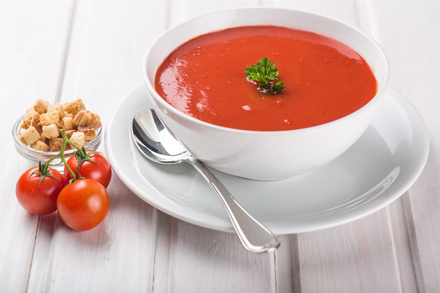 Sopa de tomate en una taza de loza blanca sobre una mesa de madera blanca.