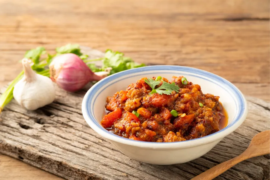 Un cuenco de nam prik ong, una salsa de chile tailandesa, sobre una superficie de madera rústica con ajo, chalotes y cilantro en el fondo.