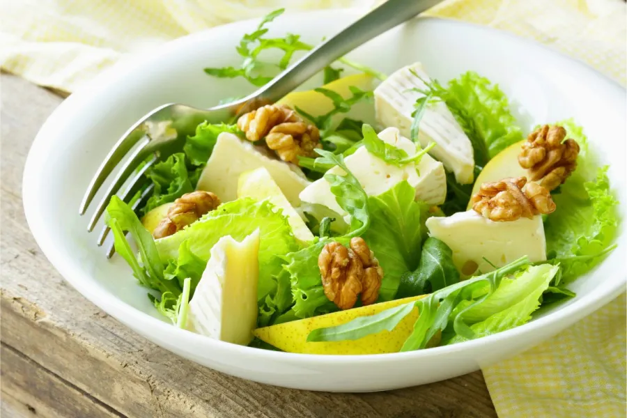 Un plato con ensalada de hojas verdes, queso camembert y pera.