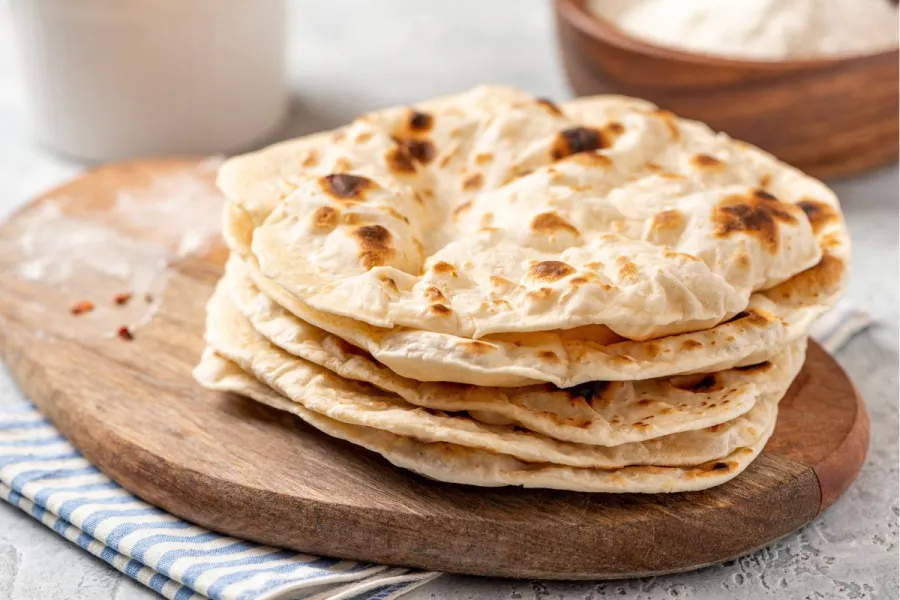 Una pila de panes planos tipo chapati recién hechos, colocados sobre una tabla de madera con un paño rayado debajo.