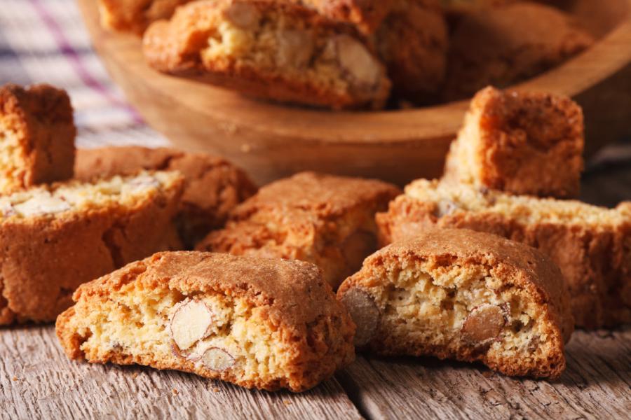 Unos biscotti de almendra sueltos sobre una mesa de madera y un recipiente de madera con más biscotti al fondo.