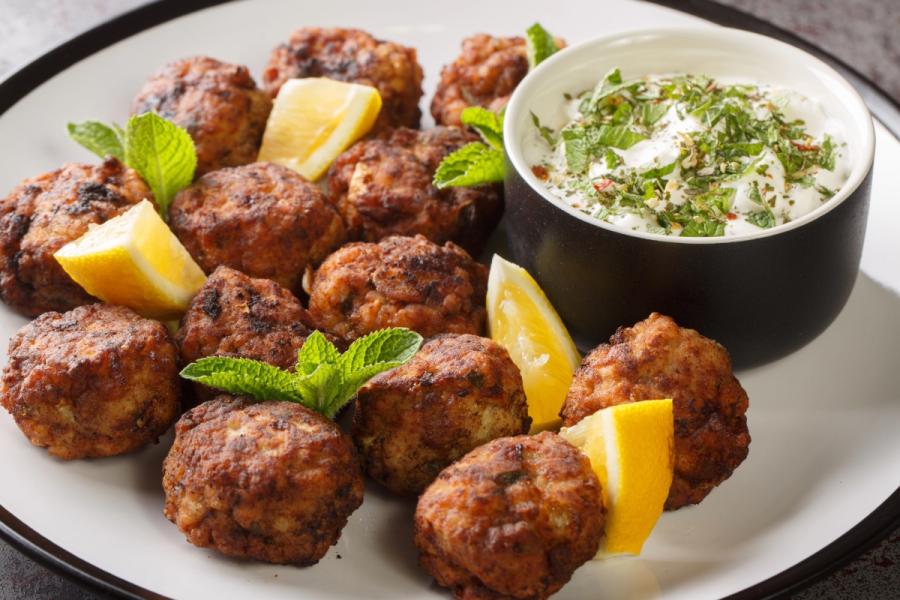 Albóndigas griegas fritas conocidas como keftedes, hechas con carne, servidas con tzatziki y trozos de limón en un plato de loza blanco.