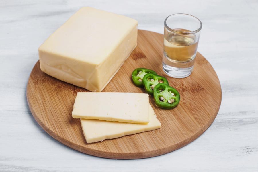 Bloque y rebanadas de queso chihuahua sobre una tabla redonda de madera, acompañados de rodajas de jalapeño y un vaso pequeño con tequila.