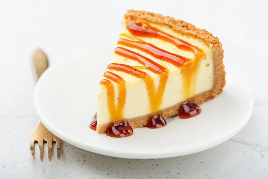Porción de tarta de queso con salsa de caramelo sobre un plato blanco, acompañada de un tenedor dorado.