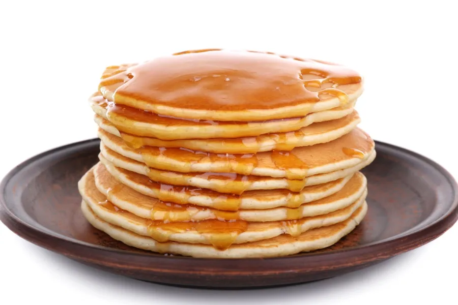 Torre de tortitas americanas doradas cubiertas con sirope de arce, servidas en un plato de cerámica oscura sobre un fondo blanco.