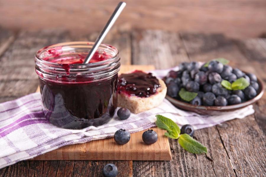 Tarro de mermelada de arándanos con una cuchara, acompañado de una rebanada de pan con mermelada, arándanos frescos y hojas de menta sobre una mesa de madera rústica.