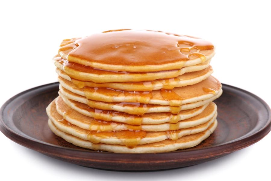 Torre de tortitas americanas doradas cubiertas con sirope de arce, servidas en un plato de cerámica oscura sobre un fondo blanco.