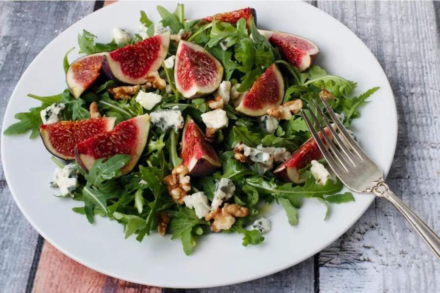 Ensalada de rúcula con higos frescos cortados en cuartos, nueces y trozos de queso azul, servida en un plato blanco con un tenedor al lado.