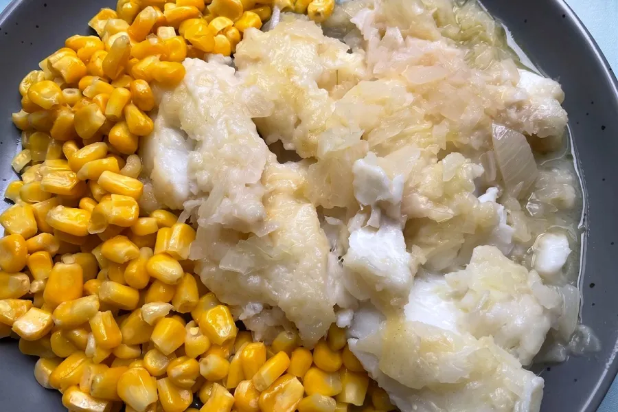 pescado blanco desmenuzado con cebolla pochada, servido junto a maíz dulce en un plato oscuro.