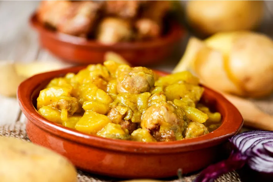 Cazuela de barro con patatas guisadas y albóndigas en salsa amarilla, con patatas crudas y cebolla morada alrededor.