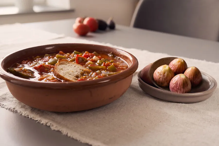 Sopa de tomate con verduras y una rebanada de pan en una cazuela de barro, con un plato de higos frescos al lado.